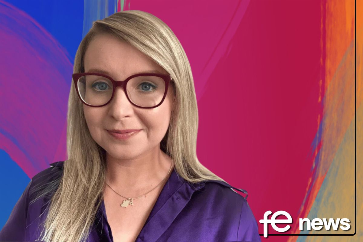 A photograph of a white woman, her head and shoulders visible as she smiles at the camera. She has long blonde hair and wears a purple shirt. Purple-tinged glasses frame her blue eyes. To her right, is a wall with blurred out, framed photographs on.
